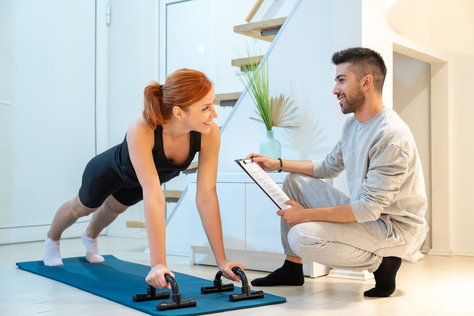 Smiling beautiful ginger woman doing home workout, push up exercise with assistance of her personal trainer at home in living room.